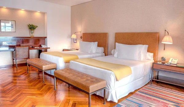 Foto de um quarto do hotel Fasano São Paulo, com duas camas com lenções branco e manda bege, onde se nota o piso em madeira e tapete.