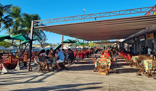 Vista de um restaurante à beira-mar com mesas coloridas, pessoas se reunindo para comer e se divertir, sob o sol em um dia claro.