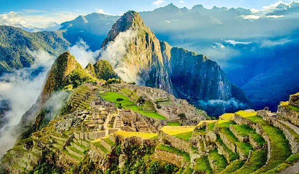 Vista panorâmica de Machu Picchu, no Peru, com antigas ruínas de pedra e extensos terraços agrícolas em meio às montanhas. Ao fundo, destaca-se uma grande montanha coberta de vegetação, parcialmente envolta por nuvens e neblina. A paisagem apresenta tons intensos de verde sob um céu azul com nuvens.