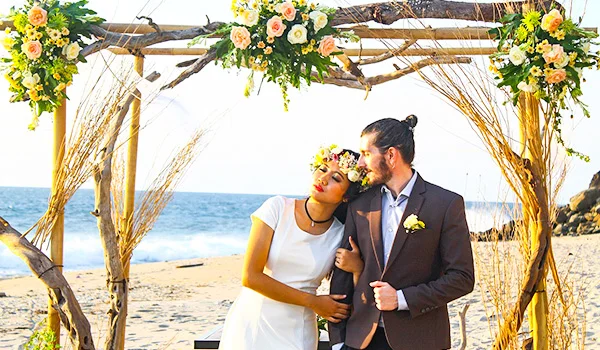 Casal de noivos em uma praia, sob um arco rústico de madeira decorado com flores amarelas, brancas e folhagens. A noiva usa vestido branco curto, coroa de flores e segura o braço do noivo, que veste terno escuro com uma flor na lapela. Os dois estão abraçados e sorrindo diante do mar, com areia e vegetação ao redor.