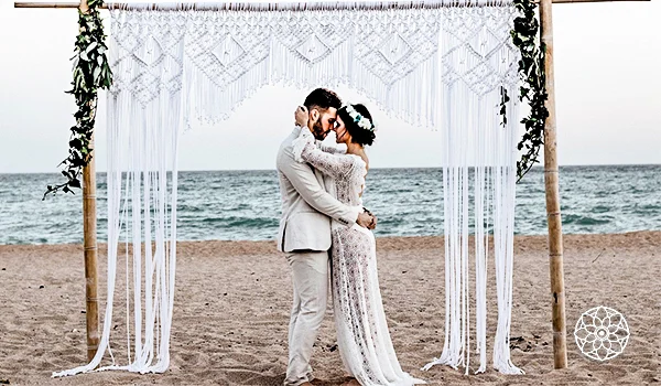Casal de noivos abraçado e se beijando em uma praia, sob um altar de madeira decorado com tecido branco rendado e folhagens. A noiva usa um vestido branco de renda e o noivo, um traje claro. Ao fundo, vê-se o mar e o horizonte. A estrutura é decorada com longas franjas de tecido branco que caem até a areia.