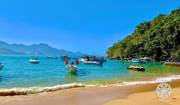 Praia em uma bela manhã de verão com águas calmas, barcos ancorados e uma vegetação exuberante ao fundo, ideal para passeios e relaxamento.