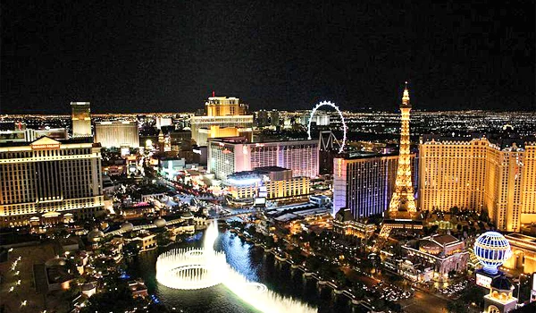 Vista aérea noturna de Las Vegas, com a região da Las Vegas Strip iluminada por milhares de luzes. Destacam-se a Torre Eiffel do Paris Las Vegas, a roda-gigante High Roller, o Bellagio e seus edifícios hoteleiros, além das fontes iluminadas do Bellagio em primeiro plano. Ao fundo, a cidade se estende sob um céu escuro.