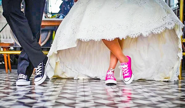 Foto de um casal de noivos mostrando apenas da cintura para baixo. A noiva usa um vestido branco de renda e tênis rosa de cano alto, enquanto o noivo veste calça social escura e tênis preto e branco. Os dois estão lado a lado sobre um piso com padrão geométrico em preto e branco.