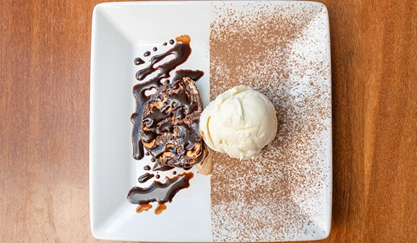 Sobremesa de sorvete com calda de chocolate, biscuit e uma generosa porção de sorvete de creme, decorada com cacau em pó em um prato retangular.