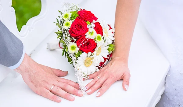 Buquê de noiva com rosas vermelhas, margaridas brancas e pequenas flores amarelas, apoiado sobre uma mesa branca. As mãos dos noivos aparecem próximas ao buquê, com a mão dele à esquerda e a dela à direita. Ao fundo, há uma cadeira branca e uma área verde desfocada.