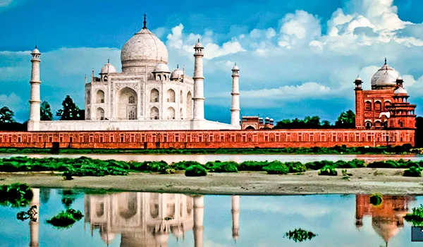 Vista do Taj Mahal, na Índia, com o grande mausoléu de mármore branco ao centro, ladeado por quatro minaretes. À direita, há uma construção histórica de tijolos avermelhados. O complexo está atrás de um muro vermelho, e um espelho d’água em primeiro plano reflete parcialmente os edifícios. Ao fundo, há vegetação e um céu azul com nuvens brancas.