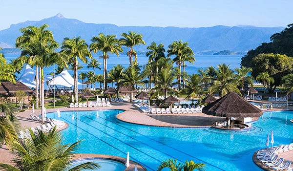 Piscina de resort em um destino paradisíaco com águas azuis cristalinas, palmeiras, bangalôs e vista para as montanhas ao fundo, ideal para férias relaxantes.