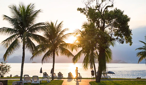 Pôr do sol na praia com coqueiros, cadeiras de praia e pessoas aproveitando o verão em uma paisagem paradisíaca.