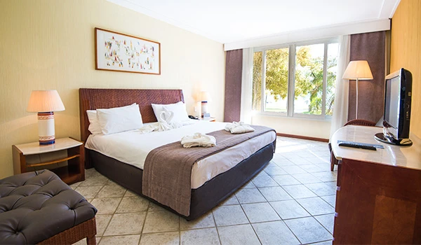 Quarto de hotel bem iluminado com cama de casal, duas luminárias ao lado e uma televisão na bancada, com janela ampla que oferece vista para árvores.