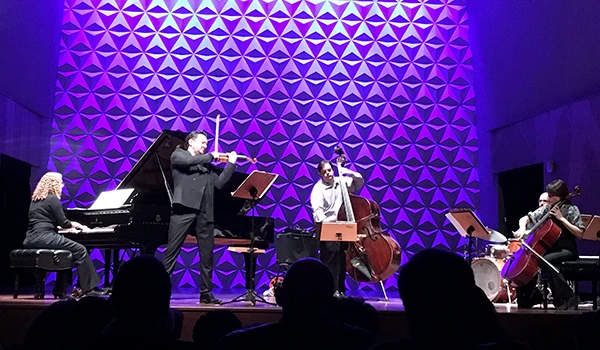 Imagem de uma performance musical de um quarteto de músicos, incluindo um violino, um violoncelo, uma pianista e um violino, em palco com iluminação azul e fundo texturizado.