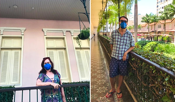 Pessoa usando máscara facial azul na rua, com parede rosa ao fundo e áreas verdes ao seu redor, demonstrando cuidado com a saúde durante a pandemia.