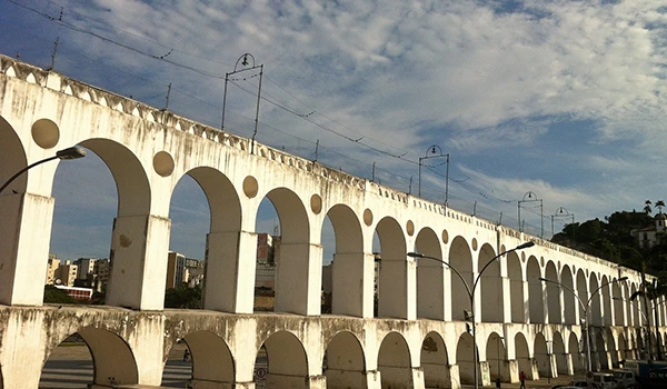 Imagem da antiga aqueduto branca com arcos, sob céu parcialmente nublado, em uma paisagem urbana, destacando a arquitetura histórica e pontos turísticos.