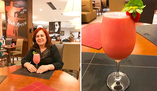 Deusa Rodrigues tomando um drinque rosa em um restaurante acolhedor, com decoração moderna e ambiente confortável ao fundo. Hotel Histórico no Rio de Janeiro.