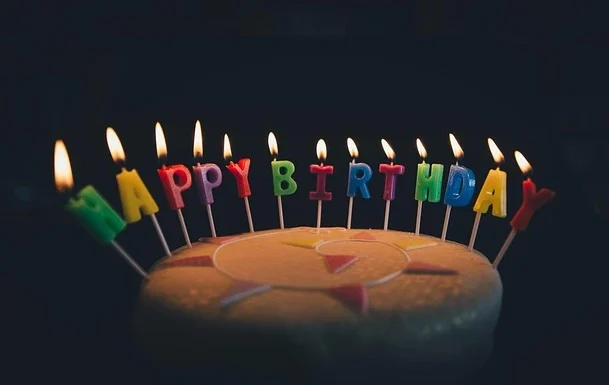 magem de um bolo de aniversário decorado com velas acesas formando a mensagem "Happy Birthday" em cores vibrantes em um fundo escuro. Perfeito para celebrações de aniversário.