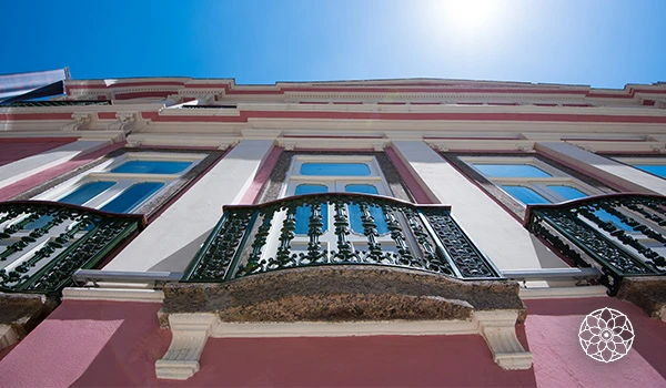 Vista de um prédio com varanda de ferro decorativa, fachada branca com detalhes rosa e janelas de vidro, sob céu azul ensolarado.
