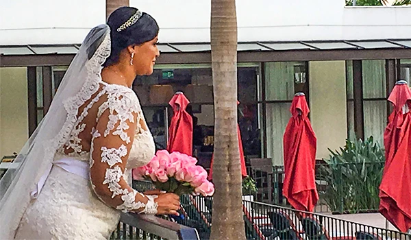 Noiva com vestido de renda attends cerimônia de casamento ao ar livre, segurando buquê de rosas rosas, cercada por decoração moderna e tropical.