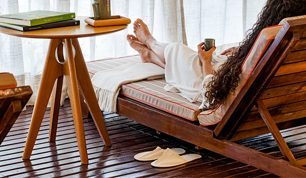 Pessoa relaxando em uma cadeira de madeira, lendo um livro e tomando chá em um ambiente confortável e elegante, com piso de madeira e cortinas brancas ao fundo.