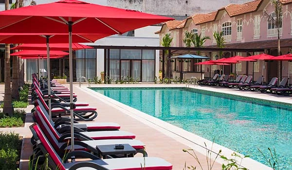 Piscina de hotel com espreguiçadeiras e guarda-sóis vermelhos ao redor, área de lazer externa bem equipada com edifícios residenciais ou comerciais ao fundo.