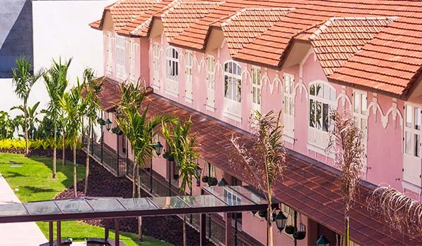 Imagem mostra um prédio com duas andares, varanda com guarda-corpos de ferro e portas decoradas, além de uma praça central com fonte e jardins bem cuidados.