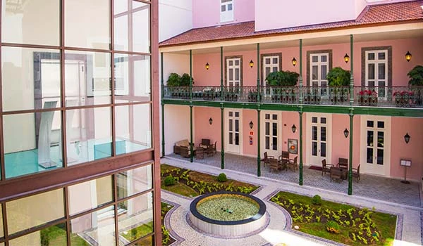Imagem mostra um prédio com duas andares, varanda com guarda-corpos de ferro e portas decoradas, além de uma praça central com fonte e jardins bem cuidados. Hotel Histórico no Rio de Janeiro.