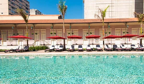 Piscina de hotel com espreguiçadeiras e guarda-sóis vermelhos ao fundo, ambiente relaxante e perfeito para aproveitar o verão no Brasil.