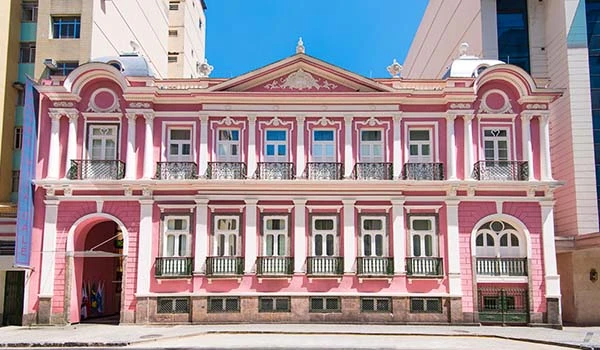 Imagem de Hotel Histórico no Rio de Janeiro, em tons de rosa com detalhes arquitetônicos clássicos localizado em uma cidade moderna, destaque para suas janelas e varandas.