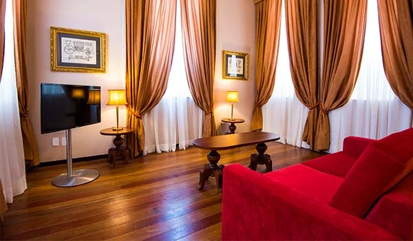 Sala de estar elegante com cortinas de cortina dourada, sofá vermelho, mesa de centro de madeira, televisão de tela plana e iluminação suave, criando um ambiente confortável e sofisticado. Hotel Histórico no Rio de Janeiro.