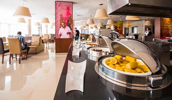 Área de café da manhã com bufê de alimentos quentes, em hotel moderno com decoração acolhedora e clientes acomodados em cadeiras e poltronas.