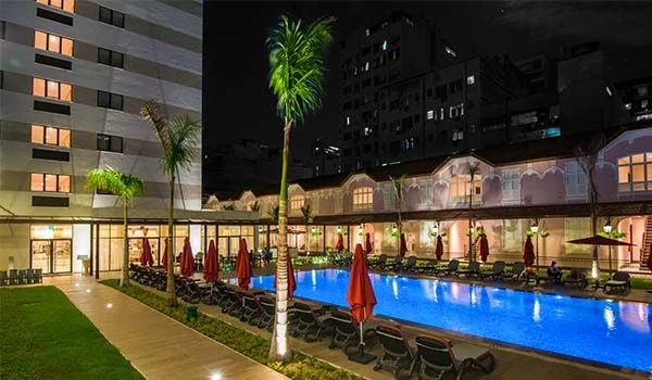 Piscina iluminada de hotel na cidade à noite, com espreguiçadeiras, guarda-sóis vermelhos e cadeiras de piscina ao redor em um ambiente sofisticado e confortável.
