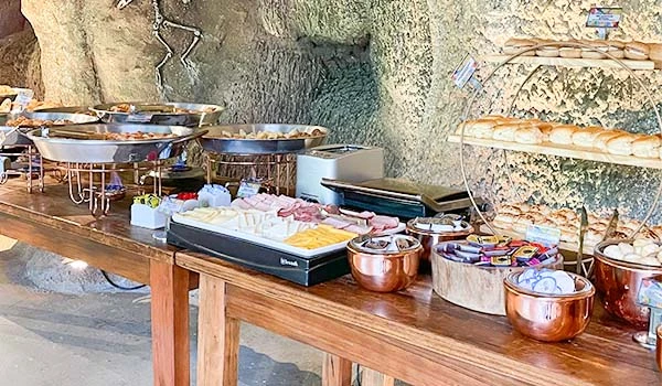 Buffet de comida variada em ambiente rústico, com pratos, saladas, queijos e acompanhamentos dispostos em uma mesa de madeira, ideal para eventos ao ar livre em contato com a natureza.