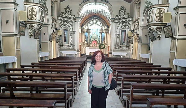 Deusa Rodrigues em interior de igreja com bancos de madeira, alta arquitetura clássica e decorativa, ambiente religioso, iluminação suave ao fundo.