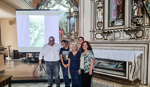 Grupo de pessoas em uma apresentação ou reunião dentro de uma igreja histórica ou antiga, com detalhes arquitetônicos elegantes ao fundo, e uma tela de projeção ao lado.