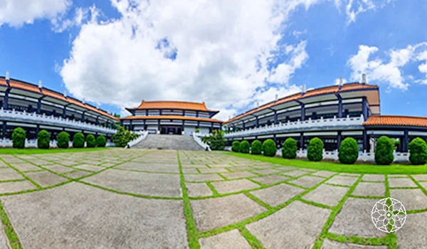 Imagem de um templo com arquitetura tradicional asiática, cercado por um jardim bem cuidado com árvores decorativas, sob um céu azul com nuvens.