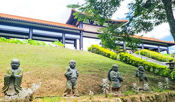 Imagem de quatro estátuas de bambu em um jardim bem cuidado, com uma casa tradicional ao fundo, cerca de vegetação e árvores, em um ambiente tranquilo e zen.