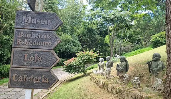 Sinalização indicando museu, banheiros, loja, cafeteria e esculturas de monjes em parque ao ar livre em um ambiente natural com árvores e plantas verdes, no Templo Zu Lai.
