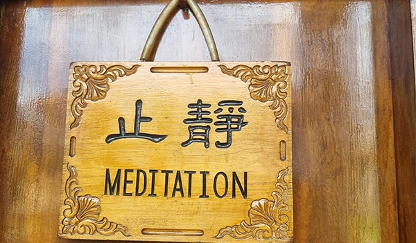 Placa de madeira com a palavra 'MEDITATION' e o símbolo chinês de silêncio, decorada com detalhes florais e suspensa por uma alça de corda, promovendo relaxamento e tranquilidade.
