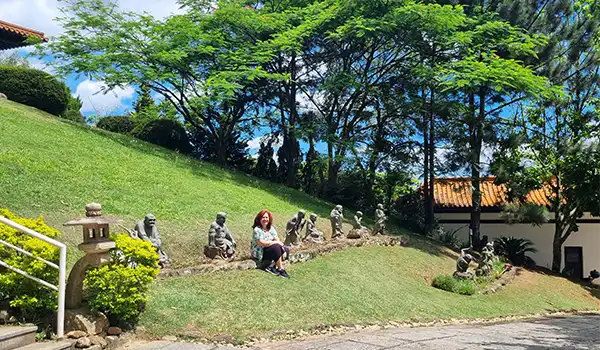 Imagem de jardins com esculturas e uma mulher sentada ao lado delas, rodeada por árvores verdes sob o céu azul em um dia ensolarado no Templo Zu Lai.