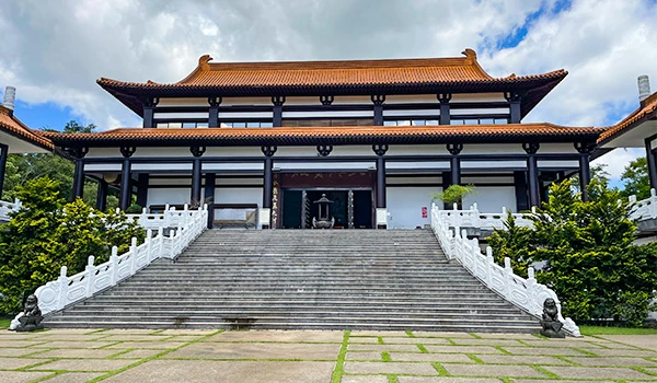 Imagem de uma tradicional construção asiática, o Templo Zu Lai, possivelmente um templo, com arquitetura de telhados avermelhados e escadaria ampla, rodeada por natureza e céu azul ao fundo.