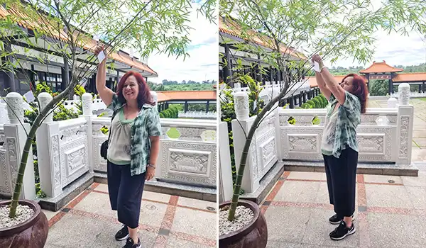 Deusa Rodrigues ao toque de uma árvore em ambiente externo, com varanda de detalhes em pedra e um jardim ao fundo, em um dia ensolarado no Templo Zu Lai.