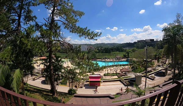 Paisagem de parque no Brasil com piscina ao ar livre, árvores verdes, céu azul com nuvens e pessoas desfrutando do espaço ao ar livre em dia ensolarado.