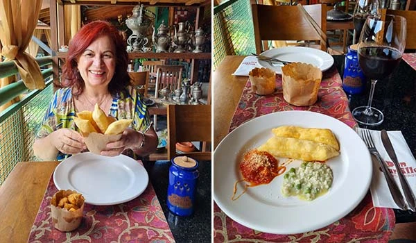 Deusa Rodrigues em um restaurante, segurando porções de pastéis, com um prato de comida ao lado e uma taça de vinho.
