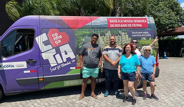 Grupo de pessoas ao lado de um veículo promocional que destaca a Basílica Nossa Senhora do Rosário de Fátima em Cotia, promovendo turismo na região.