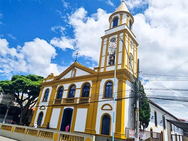 Igreja com detalhes em amarelo e branco, cercada por nuvens e céu azul, destacando a beleza arquitetônica da região.