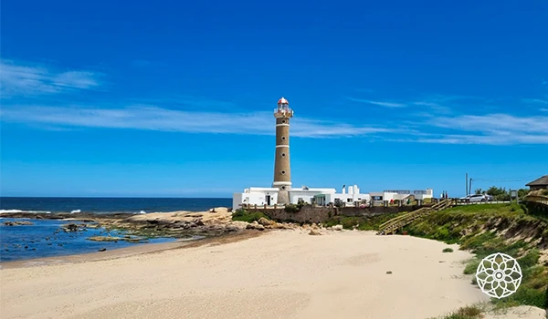 Farol na beira da praia com areia clara, rochas e oceano azul ao fundo, céu limpo e uma vista panorâmica do litoral.