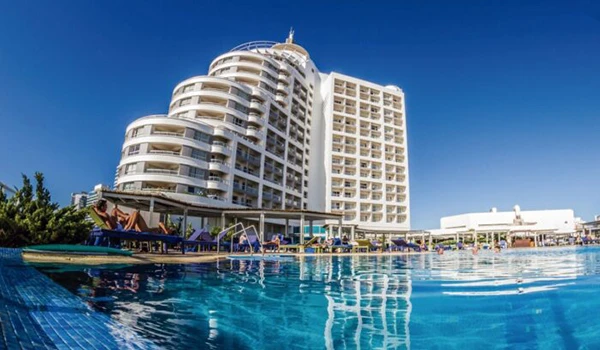 Imagem de um resort de luxo com piscina ao ar livre, prédio alto e moderno, cercado de vegetação e com céu azul claro ao fundo, ideal para férias e hospedagem de luxo.