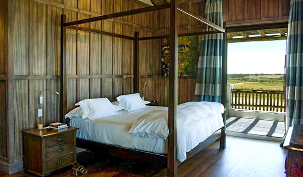 Quarto de uma acomodação rústica com cama de duas pessoas, painéis de madeira nas paredes, varanda com vista para a natureza, cortinas com listras azuis e brancas e iluminação natural.