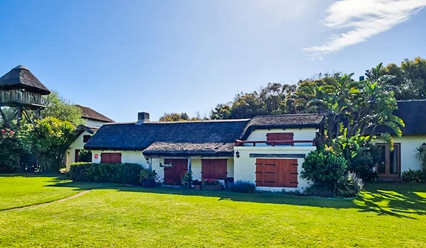 Imagem de uma casa de campo com telhado de duas águas, paredes brancas, janelas com persianas vermelhas, cercada por um jardim bem cuidado e árvores ao fundo, sob um céu claro com poucas nuvens.  José Ignacio paradores e restaurantes.
