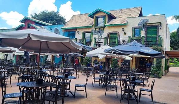 Área externa de um restaurante com mesas e cadeiras em um ambiente agradável, sombrinhas para sombra e um edifício colorido ao fundo.