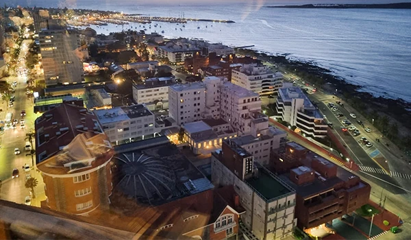Vista noturna da orla da cidade com edifícios iluminados próximo ao mar, mostrando uma praia à direita e um horizonte com céu estrelado.